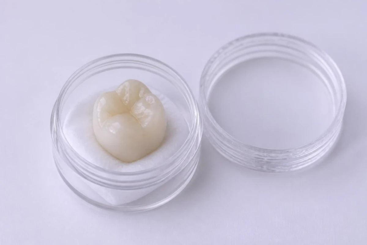 3D illustration of a dental crown standing upright on a small pad in a clear container, the chewing surface on top, with the lid beside it