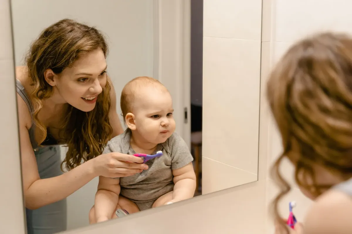 A mother smiling as she helps her baby brush teeth in front of a bathroom mirror