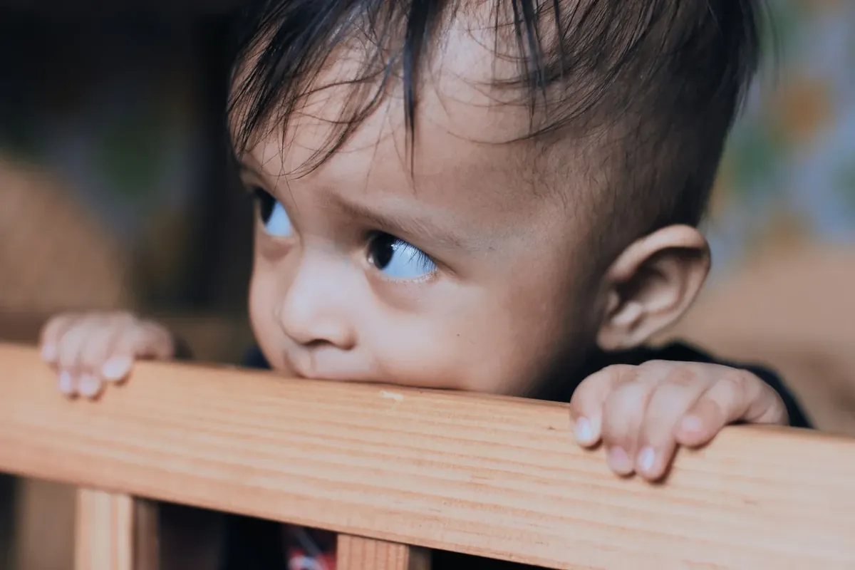 A toddler chewing on the wooden rail of a crib