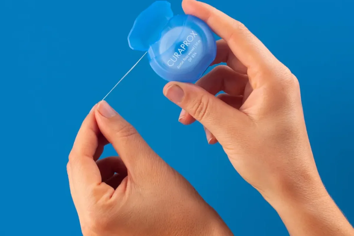 Two hands pulling a length of waxed dental floss from a round blue floss dispenser