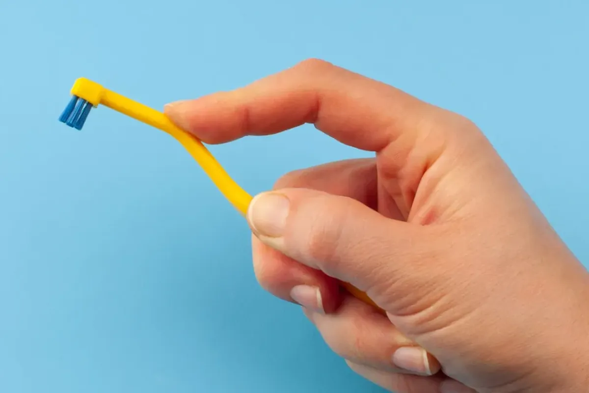 Fingers holding a yellow toothbrush with a small round head on an angled handle against a light blue background