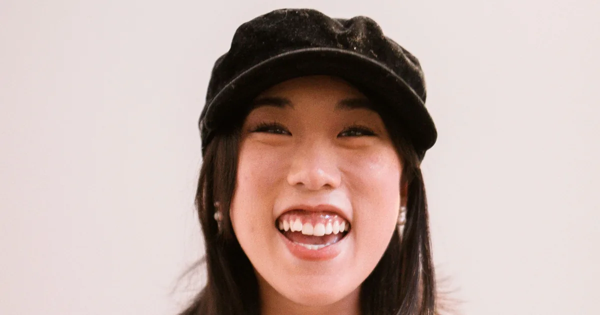 A cheerful East Asian woman in a cap laughing against a plain background