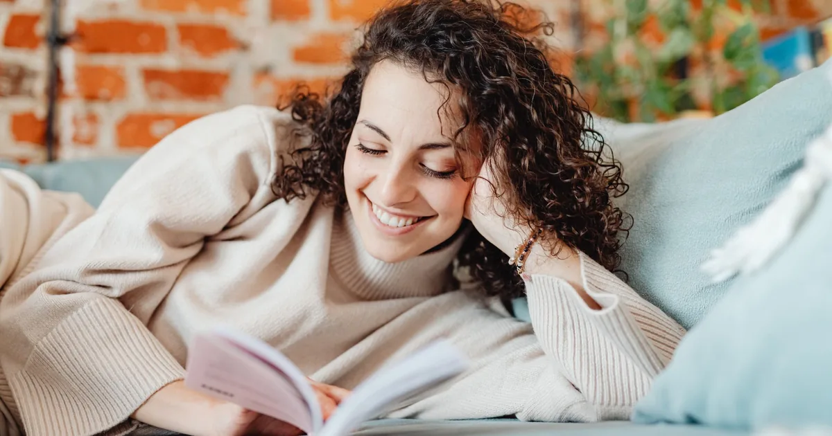 A smiling woman resting on a sofa with a pillow and reading a book at home