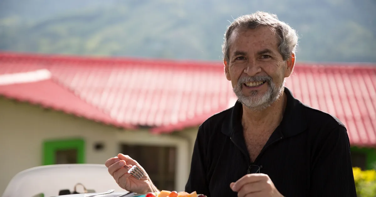 A smiling older man with a grey beard enjoying a plate of fresh fruit at an outdoor table