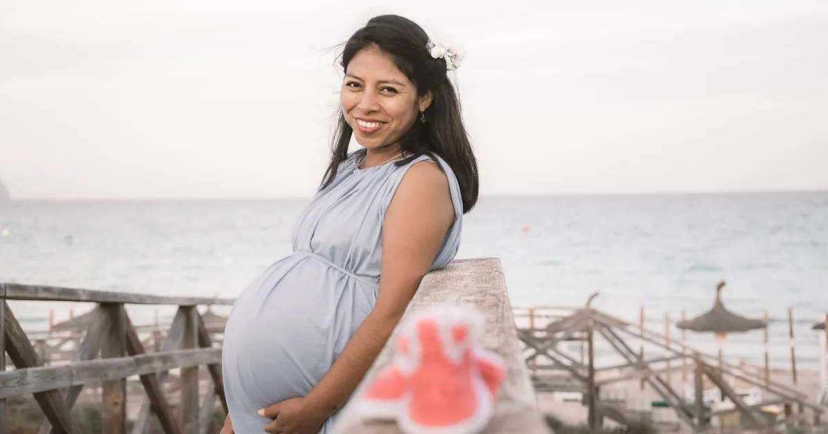 A smiling pregnant woman resting her hand on her belly by the sea