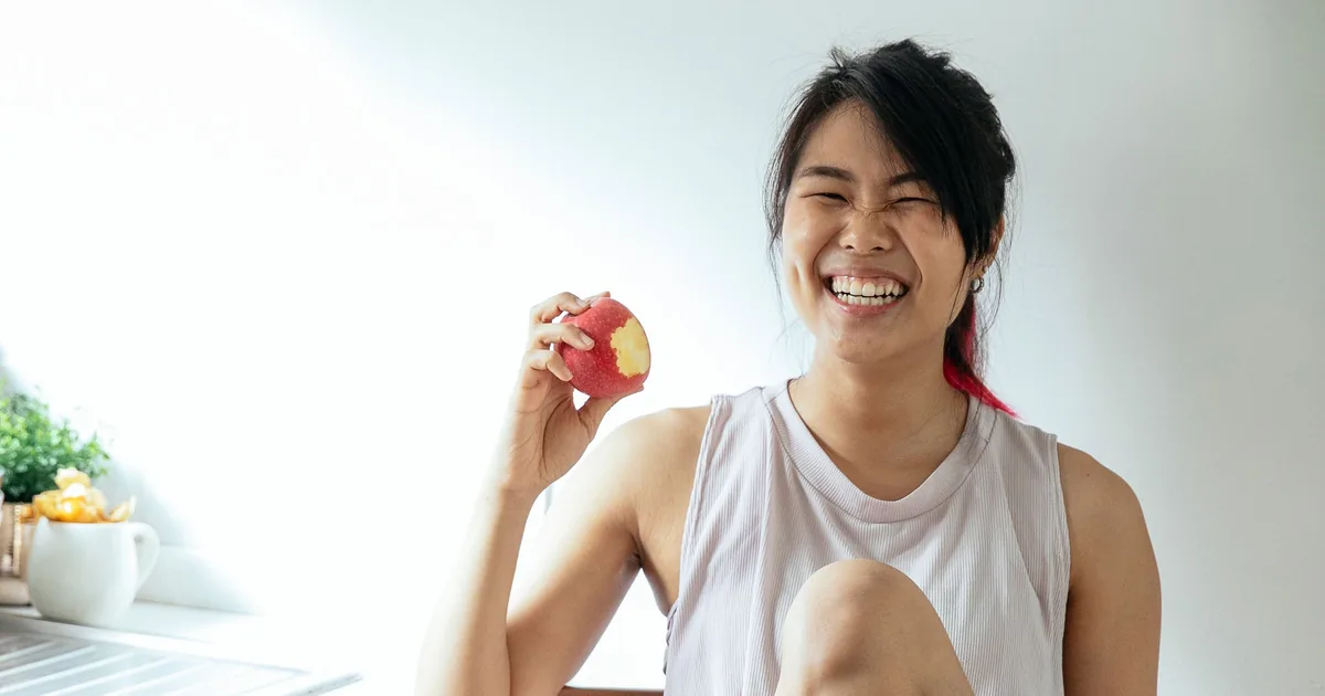 A woman laughing in her kitchen as she takes a bite of a red apple