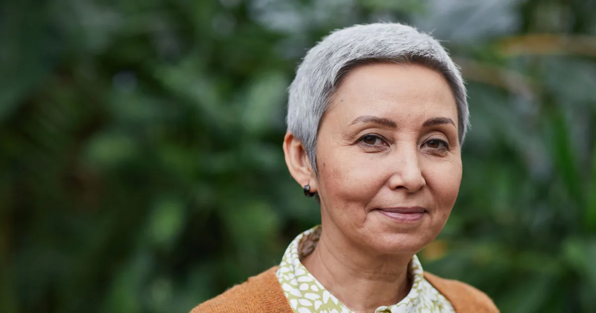A mature woman with short grey hair smiling gently outdoors among green plants