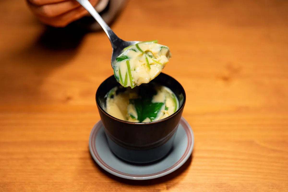 A spoonful of soft steamed egg custard lifted from a small cup on a wooden table