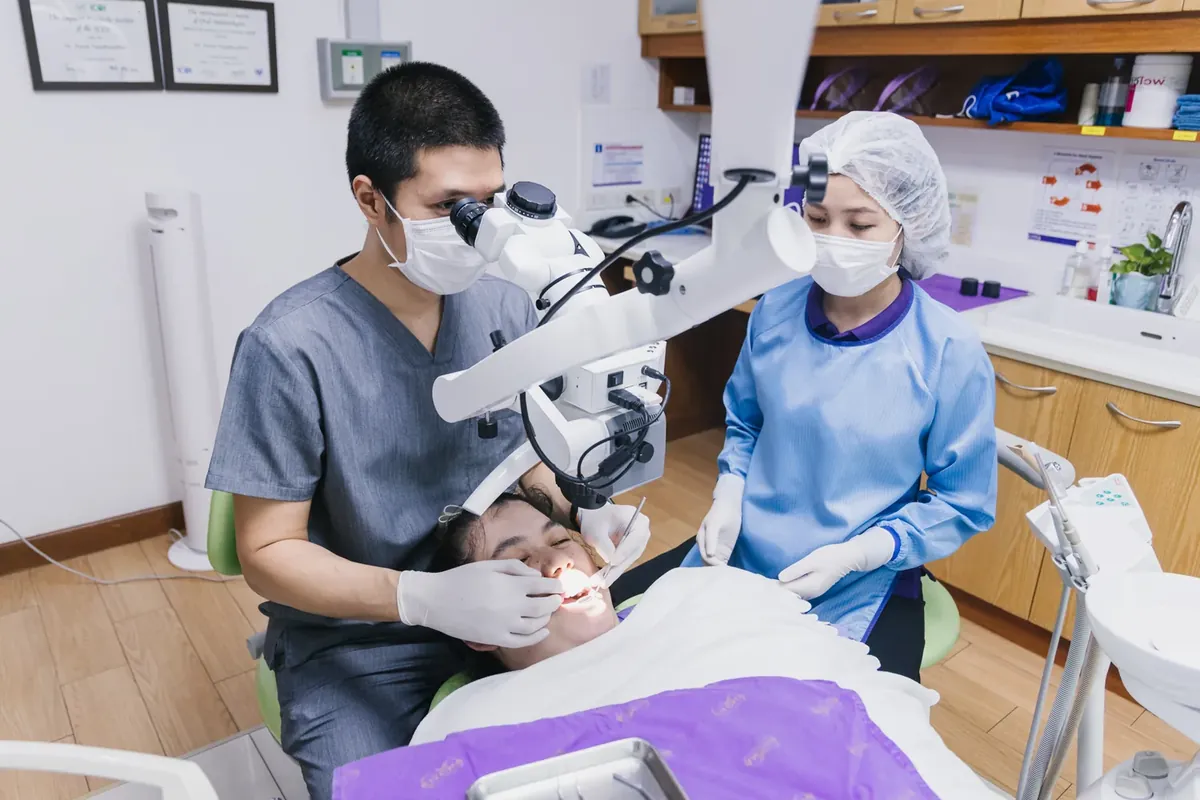 A dentist using a dental operating microscope during treatment, with an assistant beside him