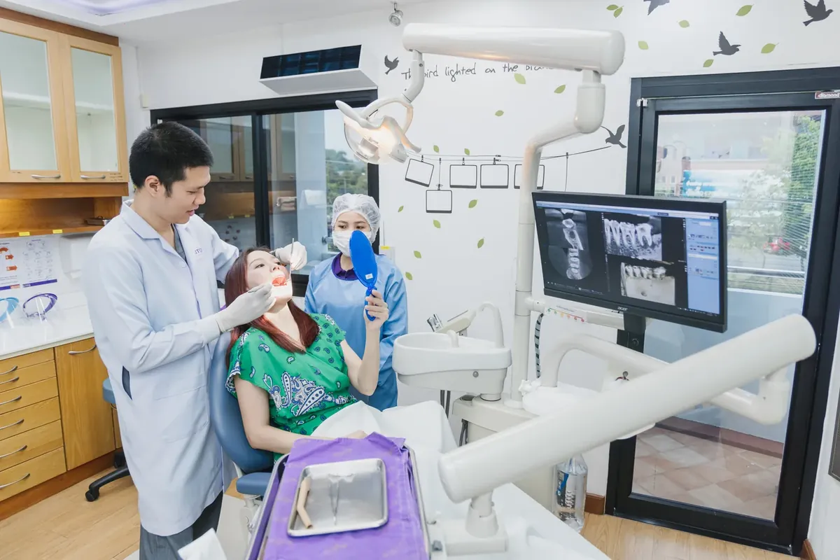 A dentist at Kitcha Dental Clinic examining a patient who holds a hand mirror, with X-ray images on the screen